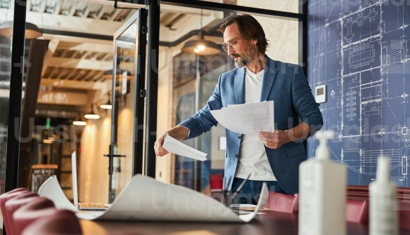 Businessman in office reviewing paper documents beside a laptop with blueprint on background wall