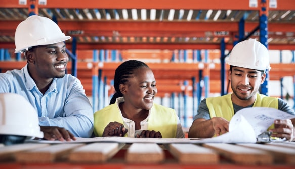 Three construction workers in hard hats smiling while reviewing documents in a warehouse