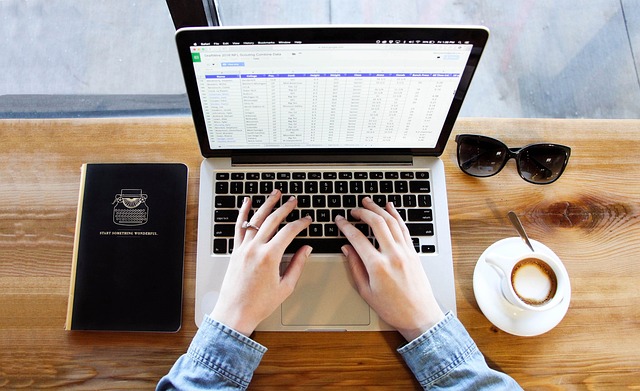 Person typing on a laptop displaying a spreadsheet, with notebook and coffee on a wooden desk