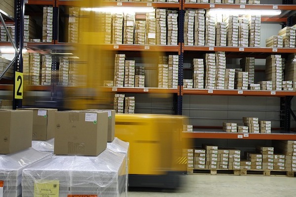 Yellow forklift moving past tall orange storage racks filled with boxes in a warehouse